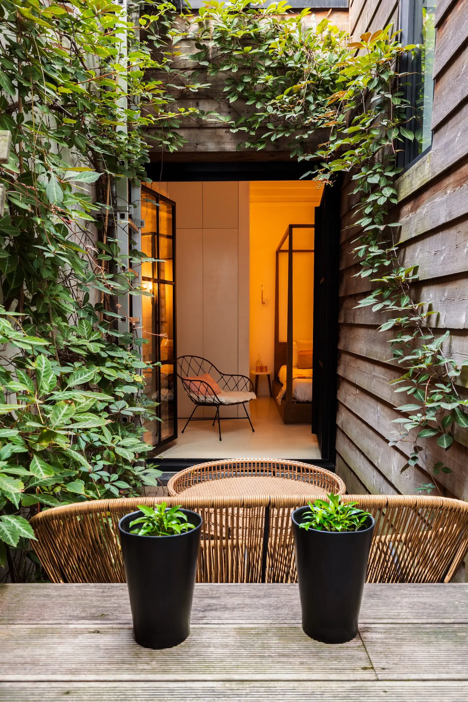 Hospitality photography of a cozy outdoor seating area with wicker chairs and a table, framed by lush green ivy and wooden walls. Through an open doorway, a glimpse of a warmly lit hotel bedroom with a four-poster bed is visible.